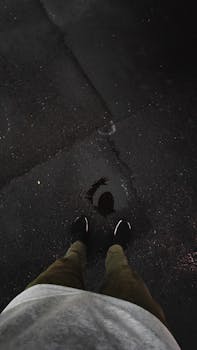 Sneakers and brown pants reflected in a puddle on a wet asphalt road, creating a moody atmosphere.
