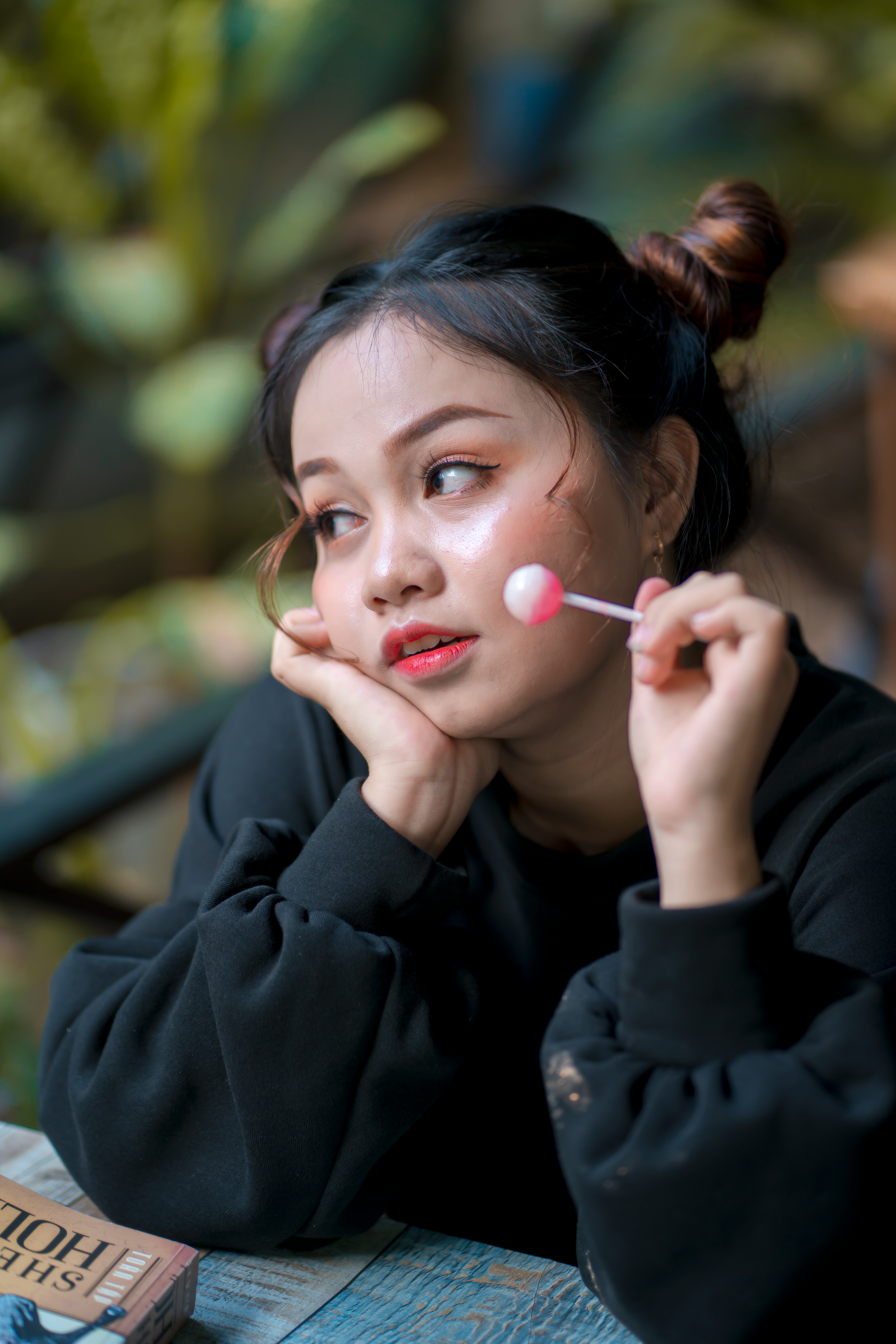 Dreamy young Asian female millennial with pigtails relaxing in cafe with hand on cheek and eating lollipop