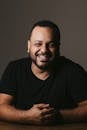 Studio shot of ethnic handsome bearded male looking at camera and smiling while sitting with crossed hands