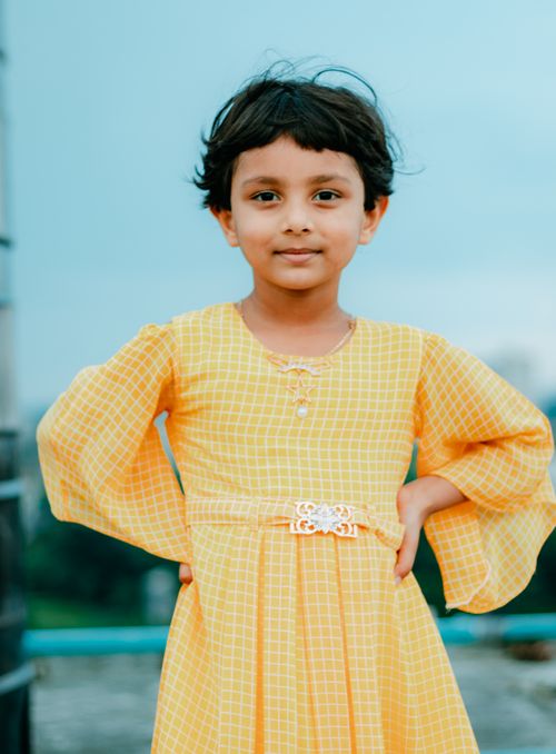 Free Smiling child in a yellow dress with hands on waist posed against a soft outdoor backdrop. Stock Photo