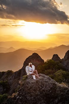 A serene photograph capturing a woman with angel wings sitting on rocky terrain during a golden sunset.