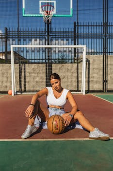 Stylish woman sitting on basketball court with ball in sunny outdoor setting.