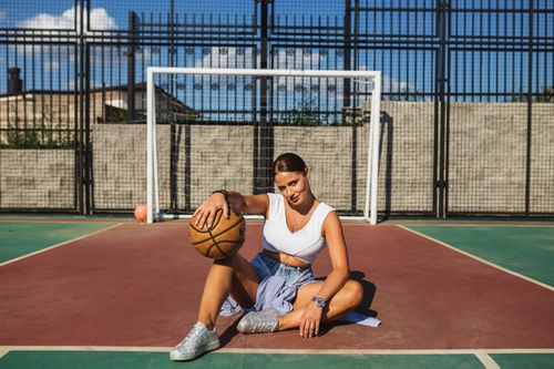 Free Young woman in fashionable outfit sitting with basketball on sunny outdoor court. Stock Photo