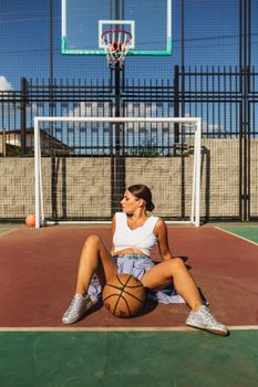 Fashionable woman in sporty attire poses with basketball on outdoor court.