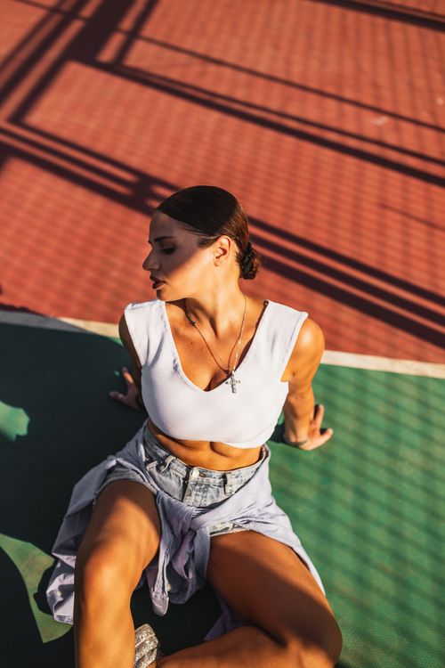Free Stylish woman in denim shorts and white tank top posing outdoors on a sunlit basketball court. Stock Photo