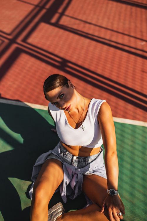 Free Model sitting on a sunlit tennis court casting geometric shadows, showcasing fashionable attire. Stock Photo