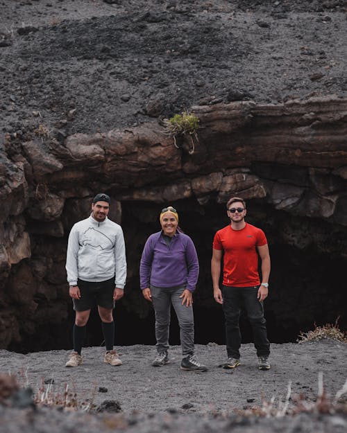 Group of multiethnic travelers in comfortable outfits standing on rocky slope near stone cave and looking at camera