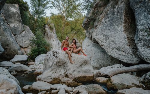Free Full body of young couple in swimsuits sitting on rocky stone in mountainous terrain and looking at camera Stock Photo