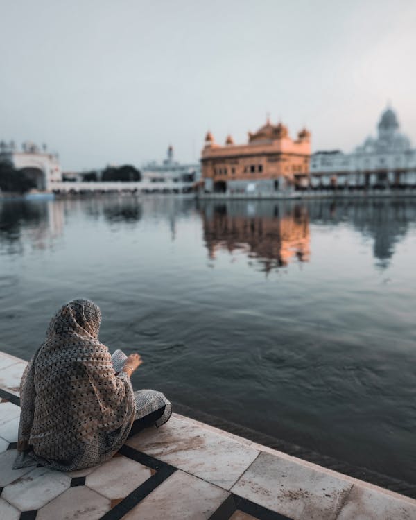 Back view of anonymous female sitting on pavement near calm lake with ancient temple on blurred background against gray sky
