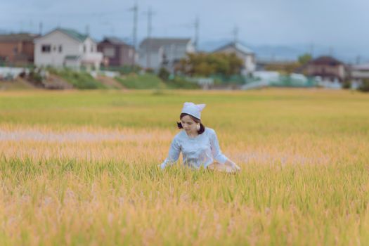 A woman in traditional attire harvesting crops in a rice field with rural houses in the background.