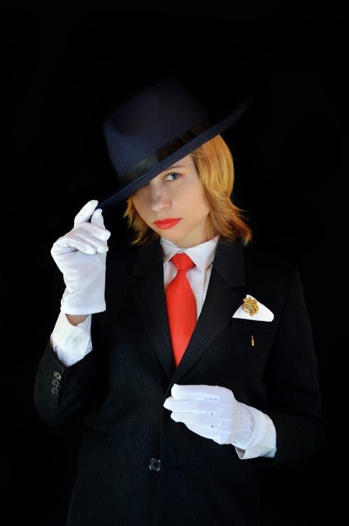Contemplative young female in classic suit and white gloves touching hat and looking at camera on black background