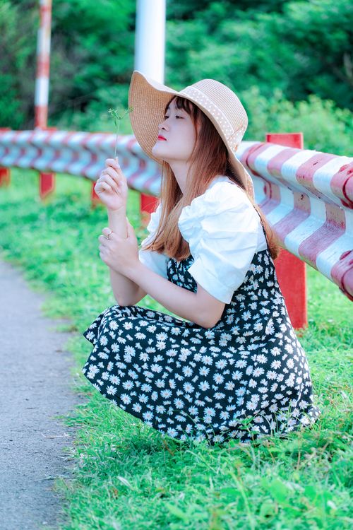 Free A young woman crouches outdoors, wearing a straw hat and floral dress, holding a flower. Stock Photo