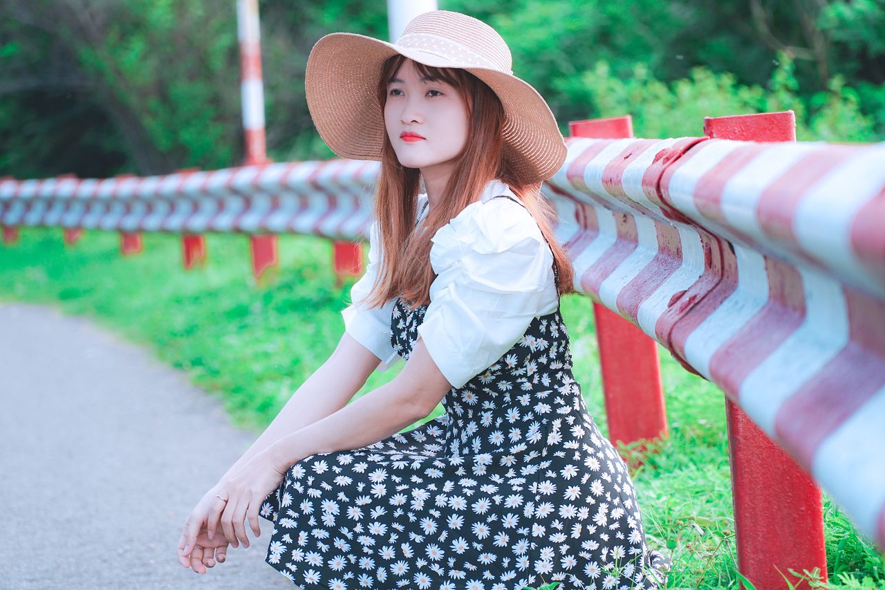 a young woman wearing a floral dress and sun hat poses by a guardrail