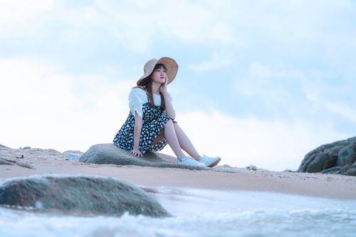 Free A young woman in a hat sits on a rock by the beach, gazing thoughtfully at the ocean. Stock Photo