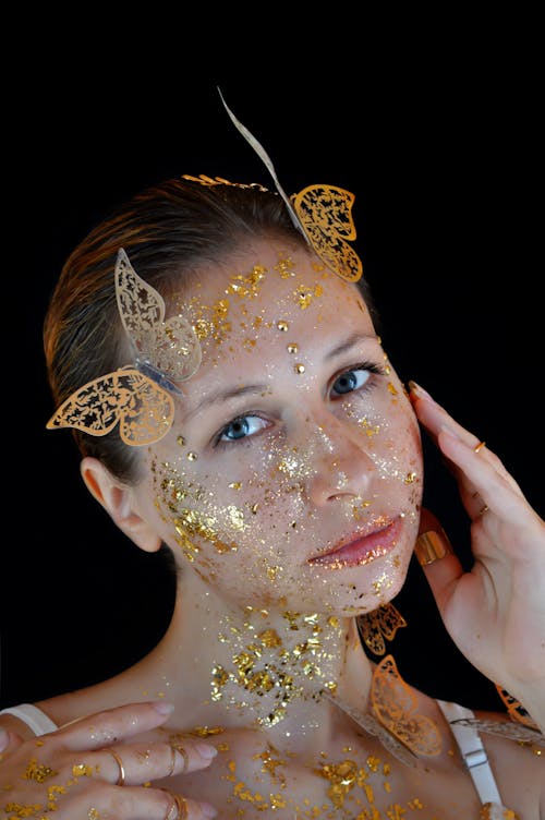 Beautiful young female model with golden shiny glitters on face and neck with artificial butterflies on head touching face and looking at camera against black background