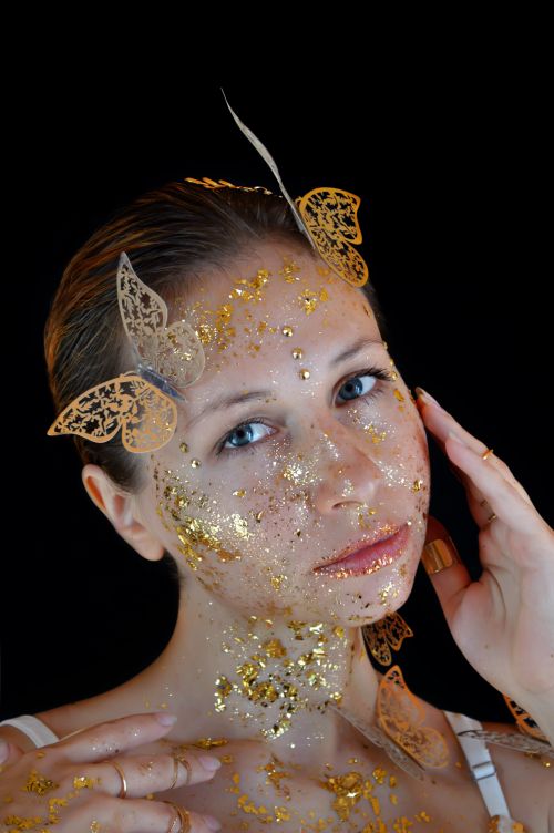 Free Beautiful young female model with golden shiny glitters on face and neck with artificial butterflies on head touching face and looking at camera against black background Stock Photo