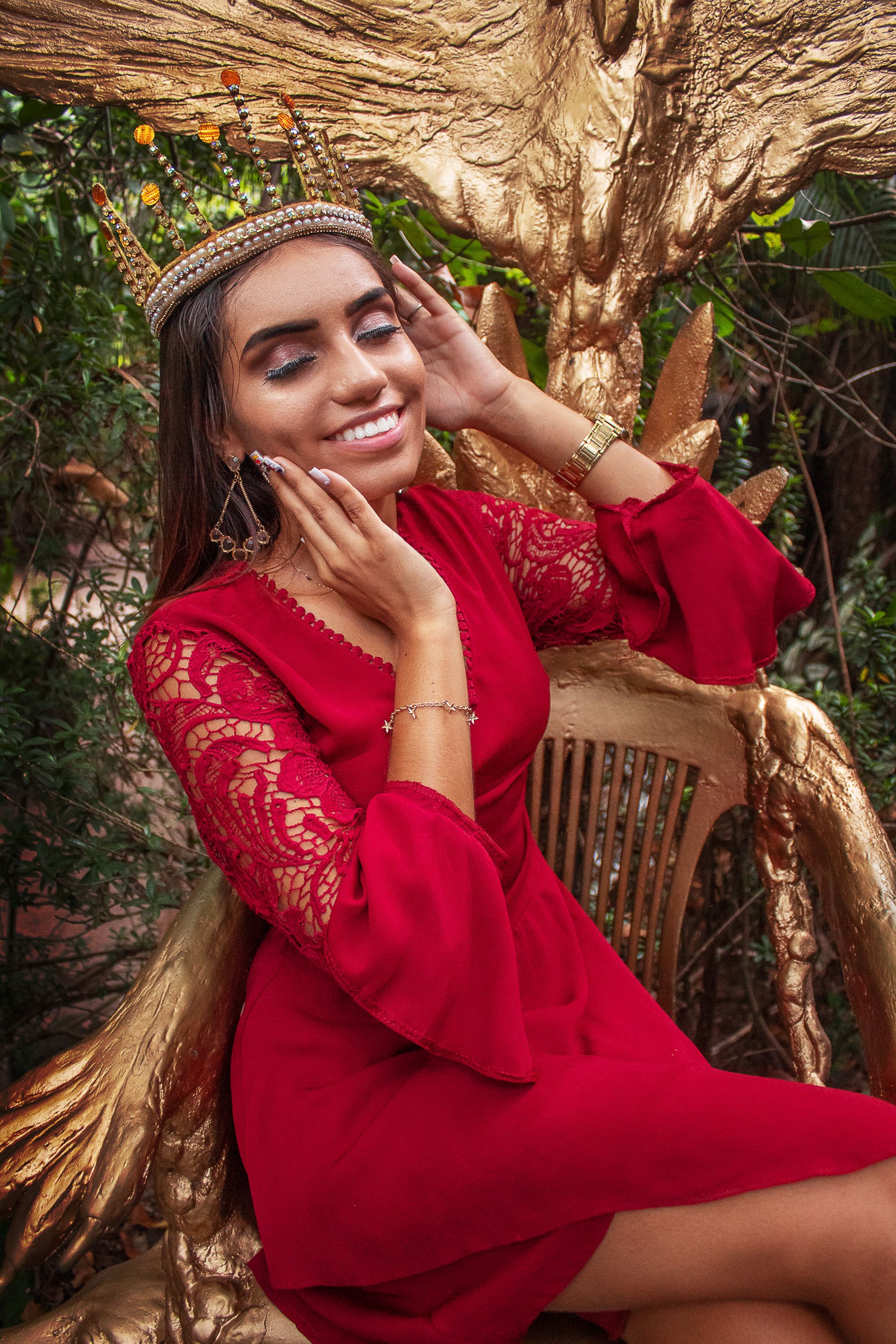 Smiling woman wearing a crown and red dress sitting outdoors, exuding elegance and fashion.