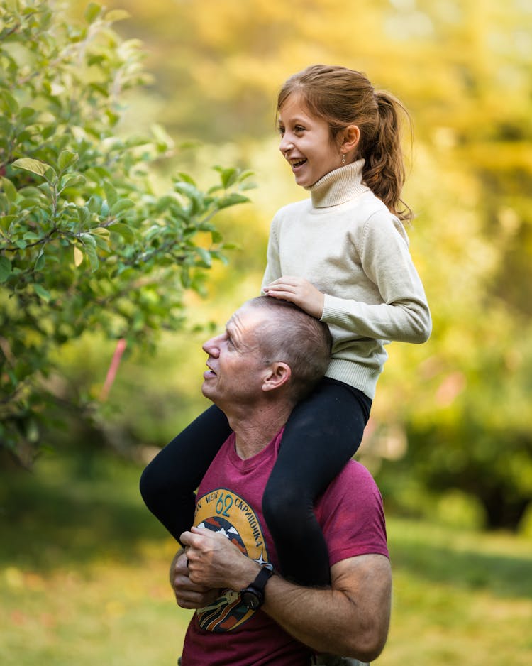 Father Carrying Cheerful Daughter On Shoulders In Lush Garden