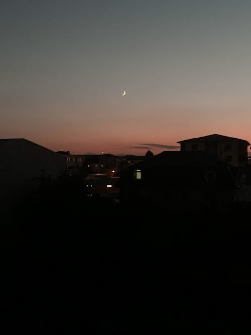 Gleaming light from windows of dark buildings located under sunset sky with shining crescent in twilight time in suburb area