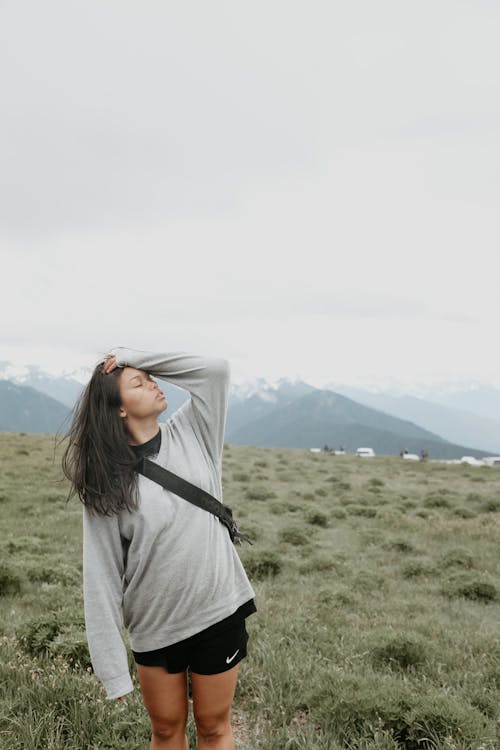 Free Charming female traveler in sportswear standing with closed eyes on green grassy hill slope against mountains Stock Photo