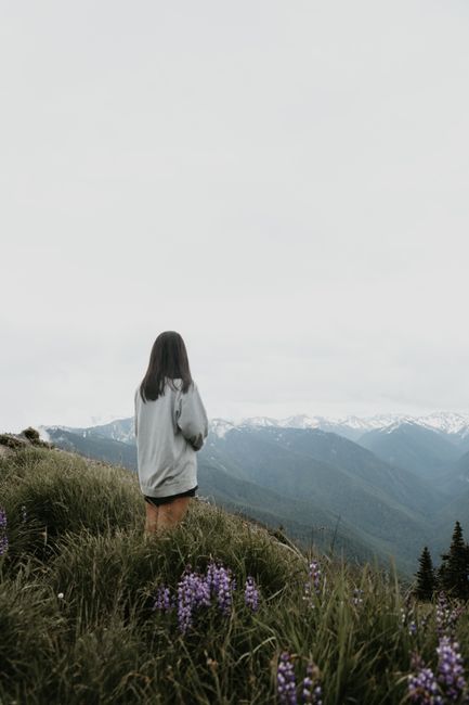 Back view of faceless female hiker standing on green grassy hill with flowers against mountains