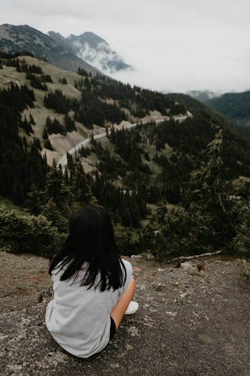 Free Back view of unrecognizable female hiker with dark hair sitting on top of mountain against green vegetate hills and clouds Stock Photo