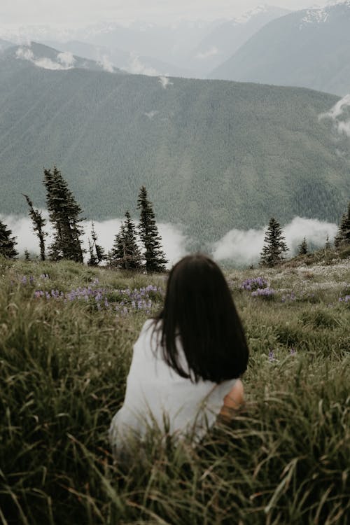 Back view of faceless female traveler sitting on grassy hill slope against mountains and clouds