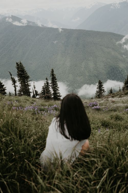 Free Back view of faceless female traveler sitting on grassy hill slope against mountains and clouds Stock Photo