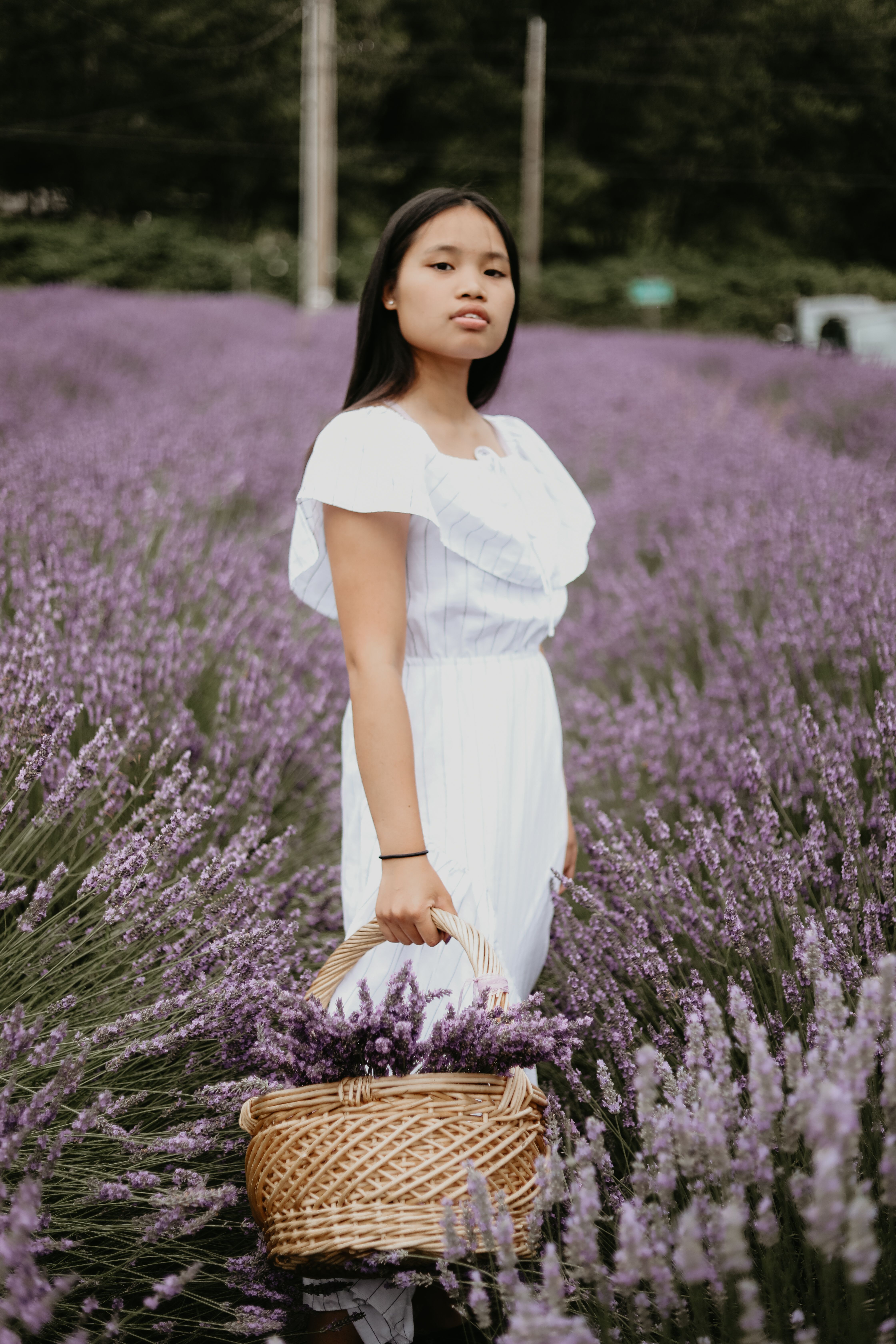 一位年轻的亚洲女性身着夏装，手提装满薰衣草的篮子，站在田野中望向镜头