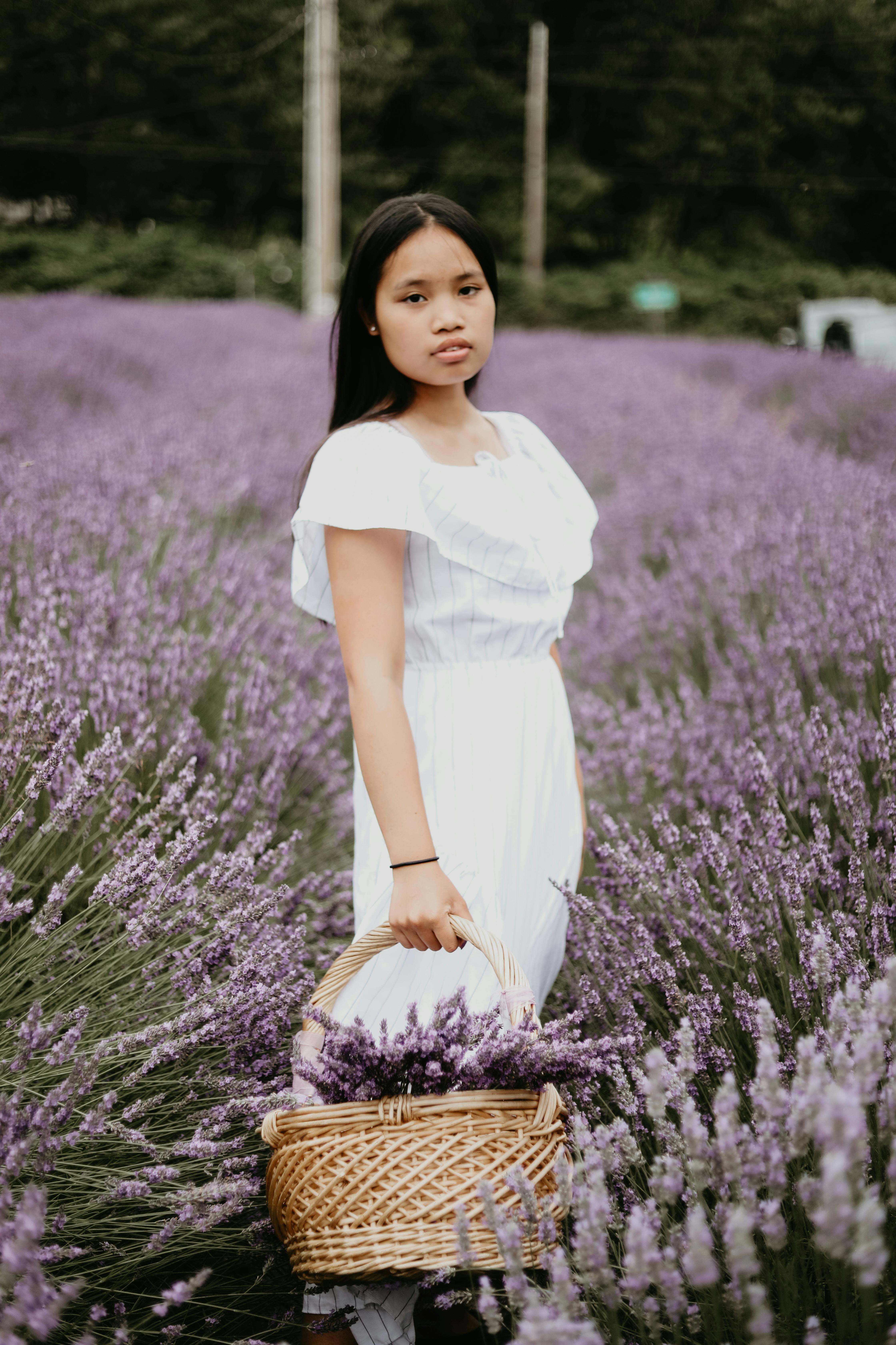 A young woman in a white dress stands in a blooming lavender field holding a basket, exuding serenity.