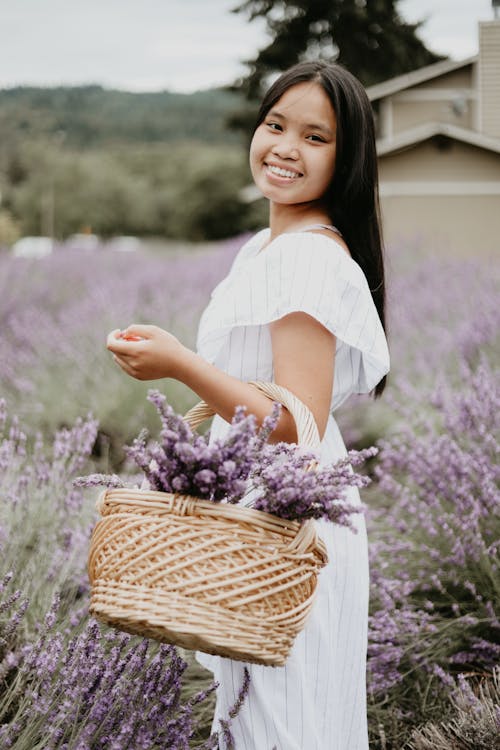 Side view of young Asian female with basket full of lavender flowers standing in meadow and looking at camera while smiling