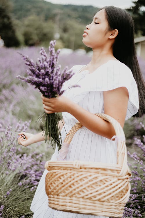 Side view glad young Asian female in white dress holding basket and bouquet of aromatic flowers on spacious field
