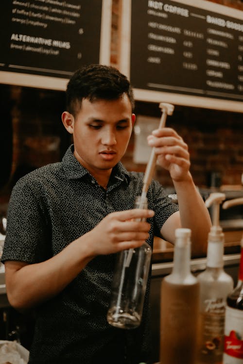 Concentrated ethnic male barista removing pump from syrup bottle while working in modern cafe