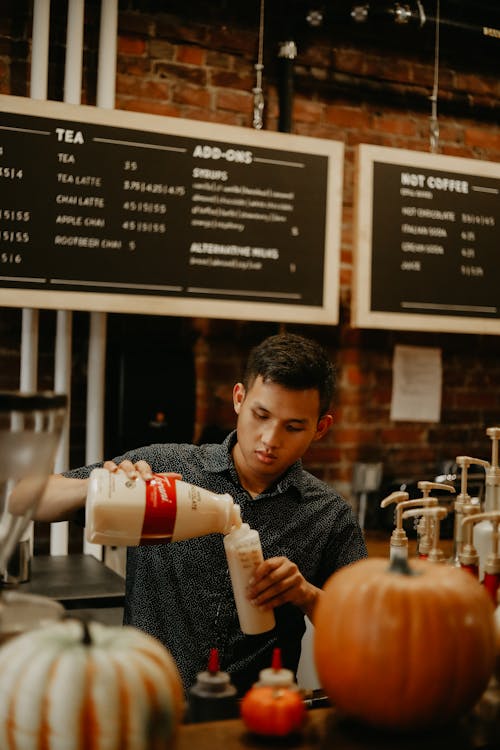 Concentrated young ethnic barista in black shirt pouring fresh milk into plastic bottle while working at modern cafeteria counter