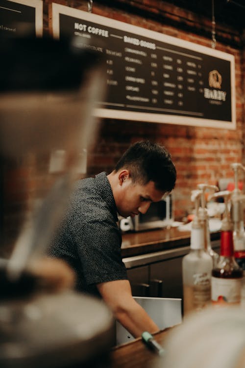 Side view focused young ethnic male barista in black uniform working at counter of contemporary popular coffee house