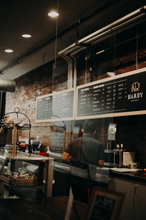 Back view anonymous male barista in black uniform working at counter behind protective glass in contemporary creative coffee shop