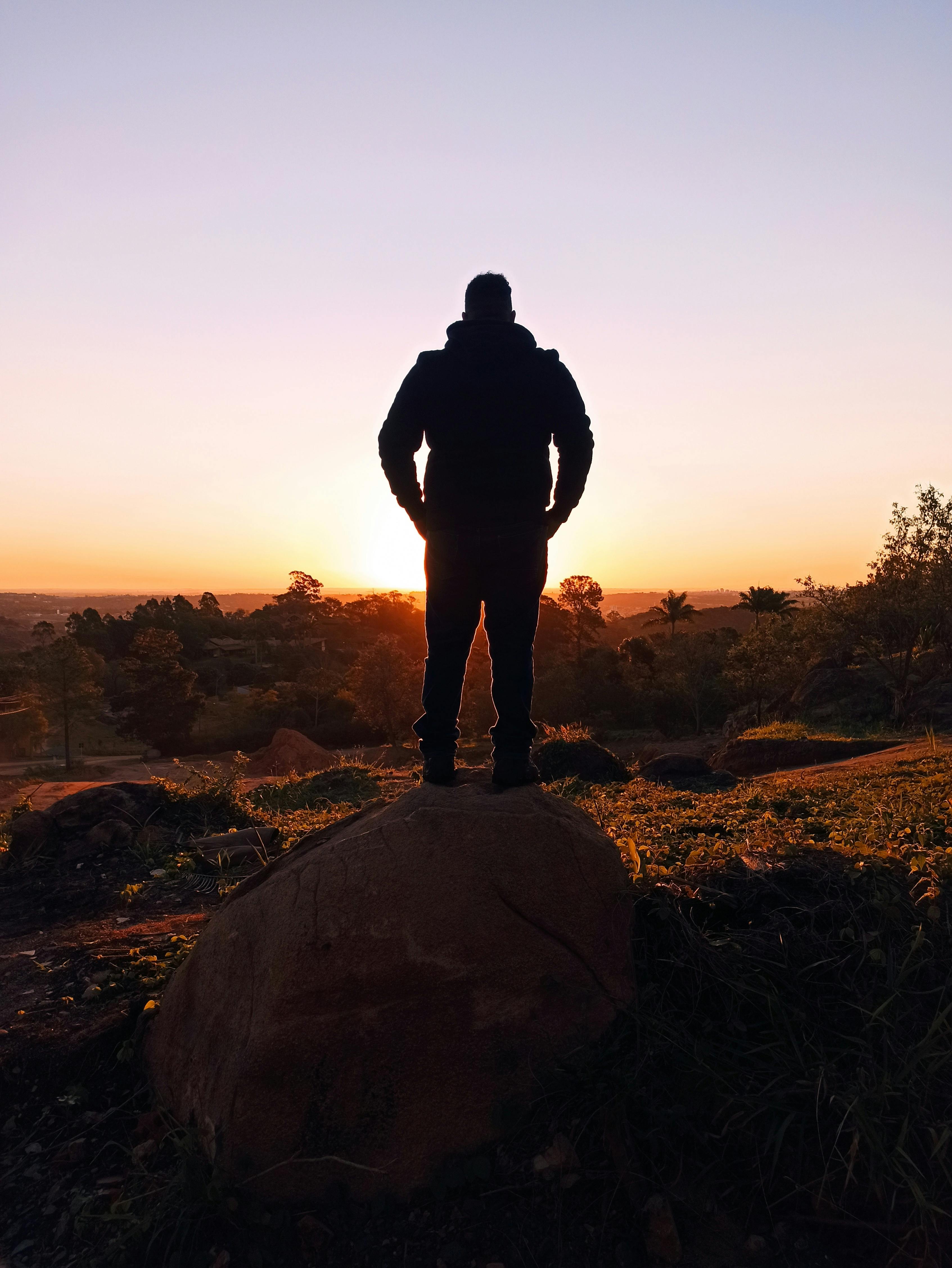 A silhouette of a person standing on a rock, facing the vibrant sunset in a natural landscape.