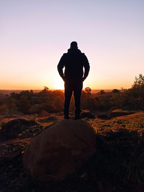 Free A silhouette of a person standing on a rock, facing the vibrant sunset in a natural landscape. Stock Photo
