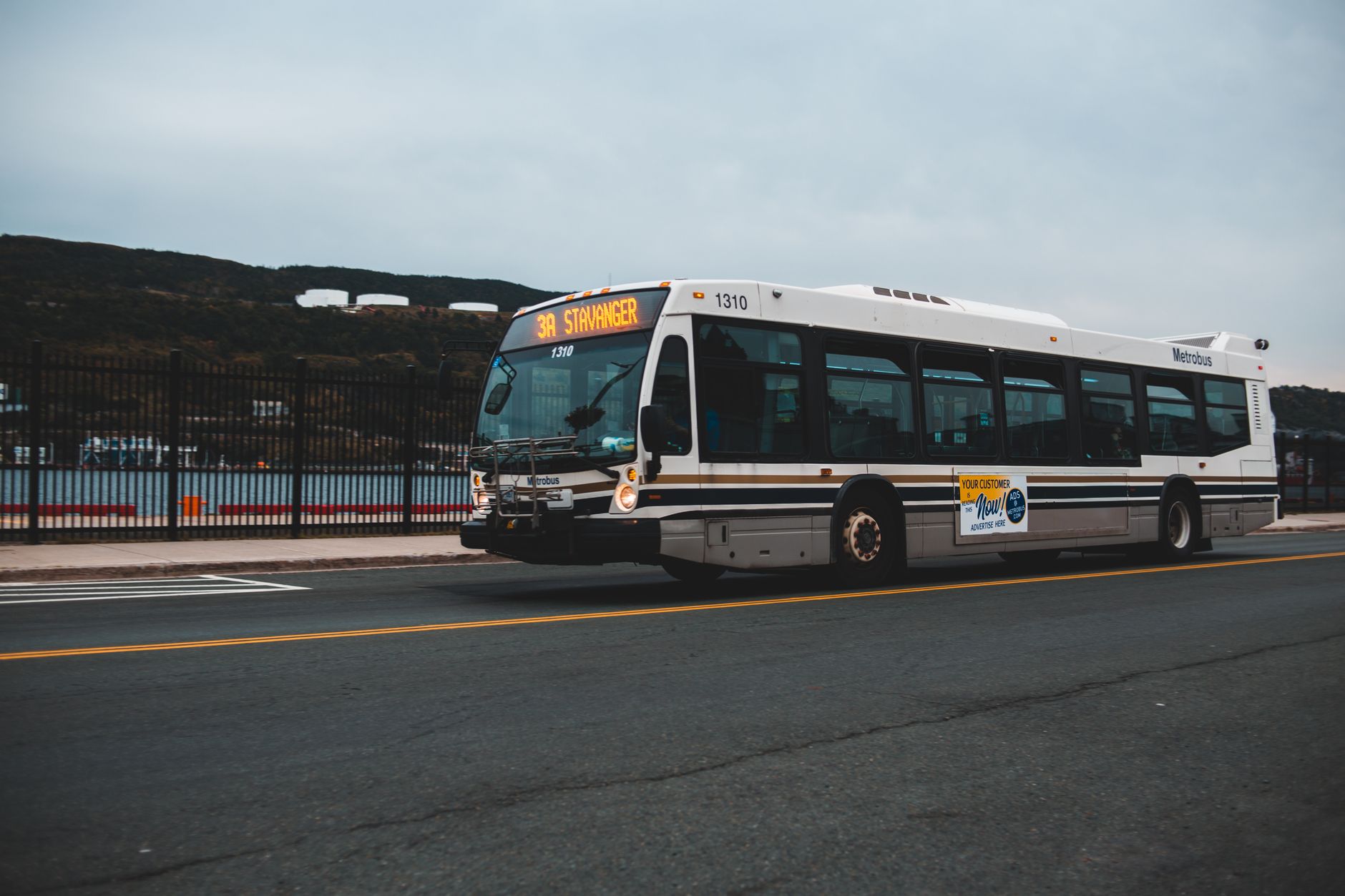Big gray bus driving on asphalt highway along metal fence near water against cloudless sky with mountains in distance outside