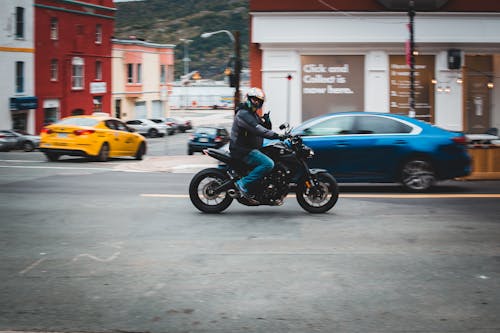 Full body of anonymous male biker wearing helmet and driving motorcycle on asphalt road with cars near residential buildings on street