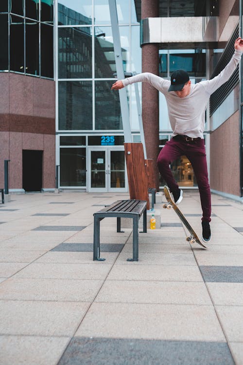 Full body of male skateboarder with arms raised jumping on skateboard near wooden bench while performing stunt on pavement in street near building