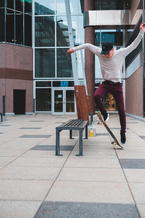 Free Full body of male skateboarder with arms raised jumping on skateboard near wooden bench while performing stunt on pavement in street near building Stock Photo
