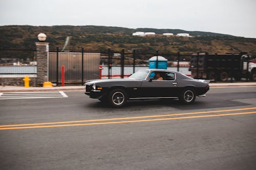 Old timer black automobile driving on asphalt roadway along metal fence near sea with hilly terrain in distance in street