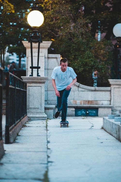 Free Full body of male skater riding skateboard on narrow pavement near metal fence with streetlight and green trees in city street Stock Photo
