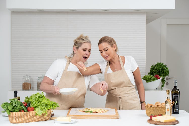 Women Wearing Brown Aprons Sprinkling Cheese On A Pizza