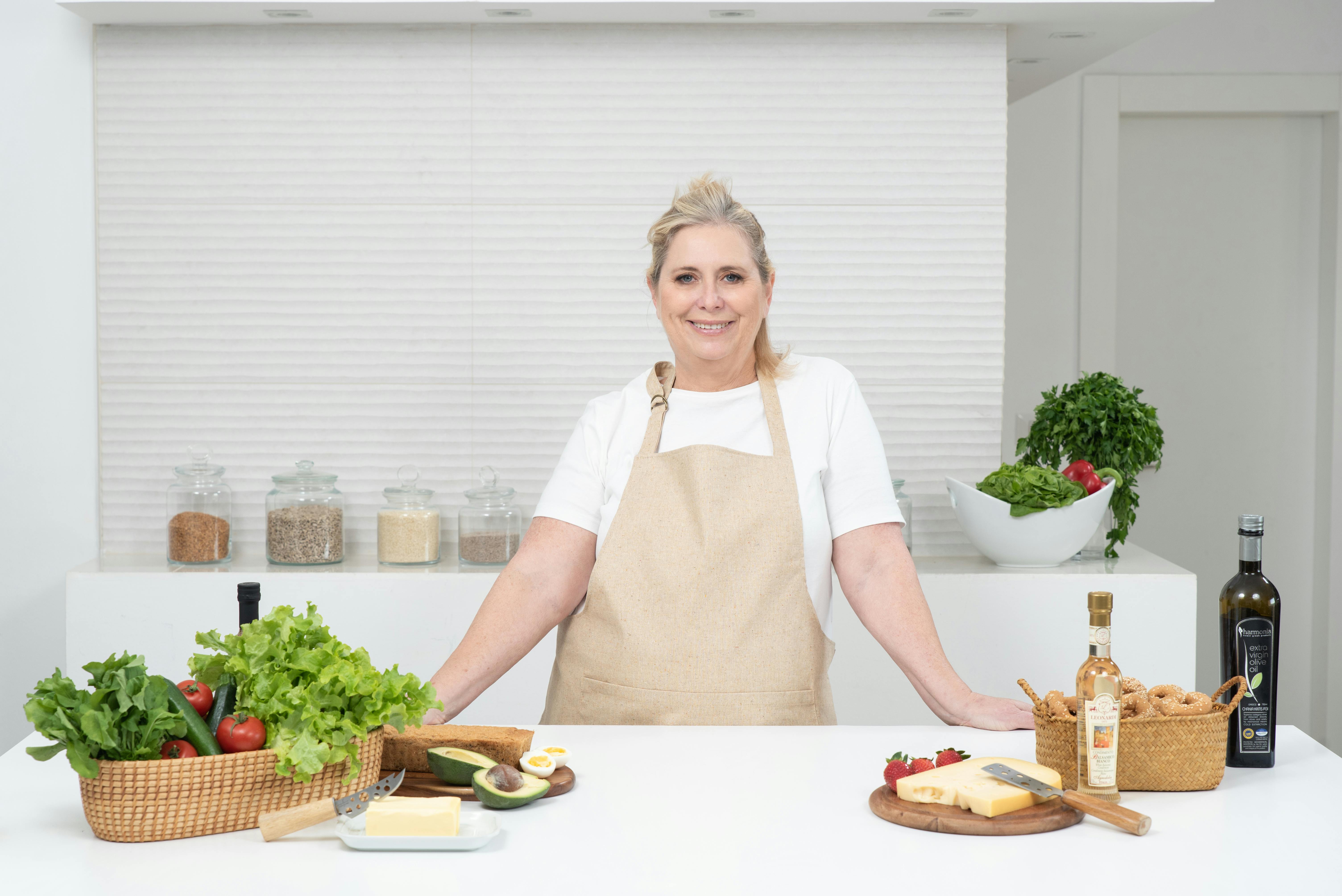 A mature woman in an apron stands at a kitchen counter with fresh produce and cheese, ready for cooking.