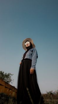 Fashionable woman in hijab and denim jacket with hat poses against a clear blue sky.