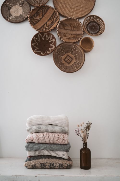 Free Neatly folded stack of sweaters against woven wall decor in a minimalist setting. Stock Photo