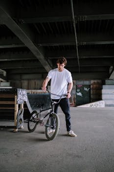 A man in a white t-shirt holds a BMX bike under a bridge in an urban setting.