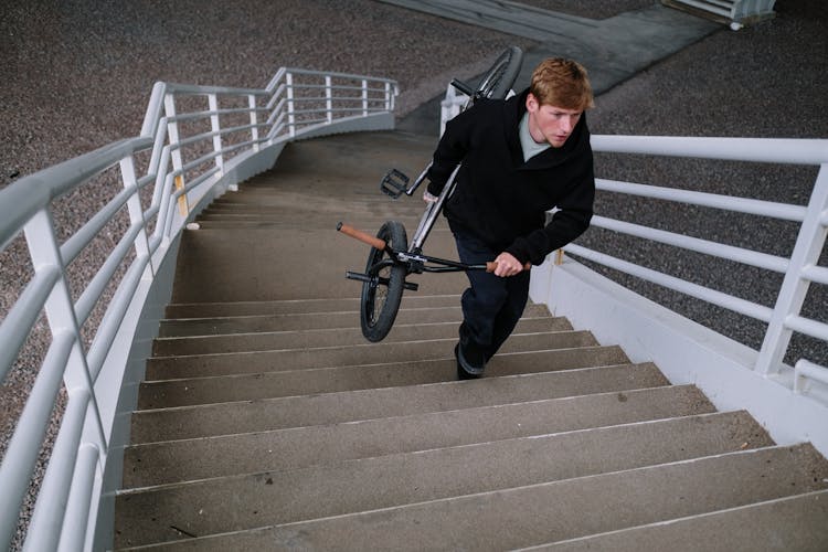 Man In Black Jacket Carrying A Bicycle While Walking Up On Stairs 